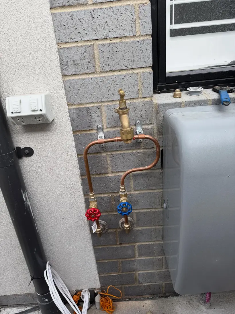 Copper pipework with a brass tempering valve above red and blue isolation valves, mounted on a brick wall beside a hot water unit