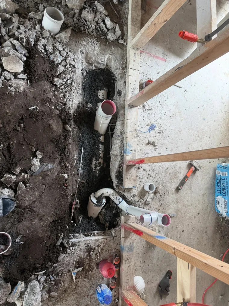Excavated drainage rough-in with white PVC pipe and junctions laid on a gravel bed beside timber formwork