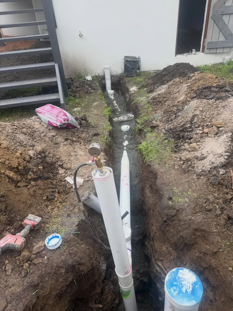 Long open trench with sewer pipework laid on a gravel bed alongside a rendered wall